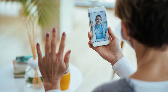 Close-up of woman greeting her doctor while using smart phone and having video call.  Focus is on female doctor on touchscreen.